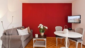 A modern living room with a red accent wall, a grey sofa, a white coffee table, a white dining table with chairs, and a flat-screen TV mounted on the wall. - Double Room by Interhome (Ascona)
