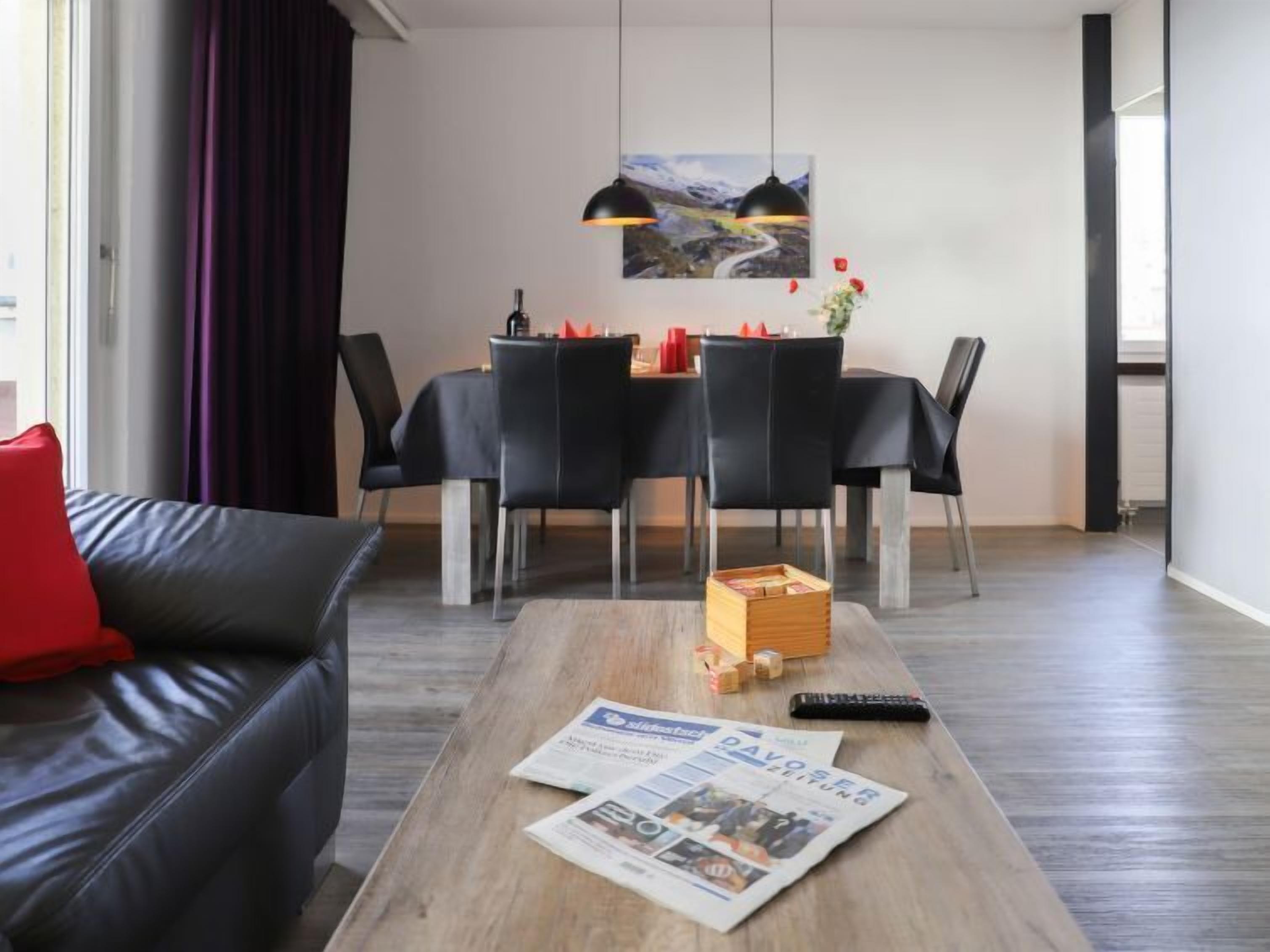 Una sala moderna con un sofá de cuero negro, una mesa de centro de madera con periódicos, una zona de comedor con sillas negras y una mesa, y una lámpara colgante sobre la mesa.