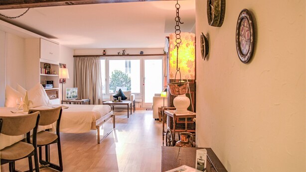 A hotel room with a bed, a desk, a chair, a table, and a window with a view of trees. - Residenz Surlej 6 by Interhome (St. Moritz)
