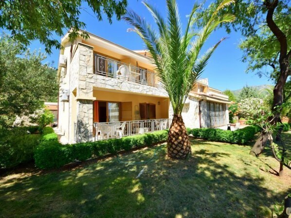 Une maison à deux étages avec une façade en pierre, un balcon doté de balustrades blanches et un jardin bien entretenu agrémenté d’un palmier.