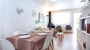 A dining area with a striped tablecloth, white chairs, a sofa, and a fireplace.