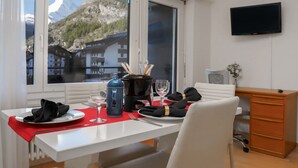 Ein Essbereich mit Tisch für zwei Personen, einer Kaffeekanne, Weingläsern und Blick auf Berge durch große Fenster.