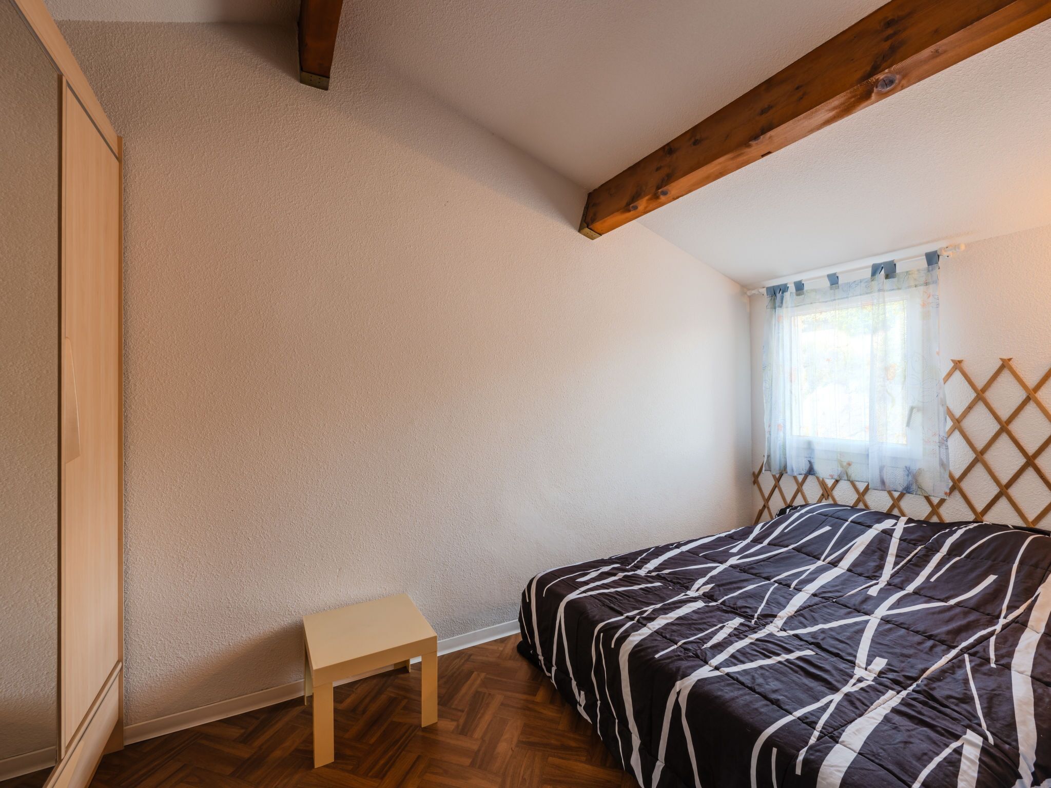 A bedroom with a bed, a small table, wooden beams on the ceiling, and a window with a curtain.