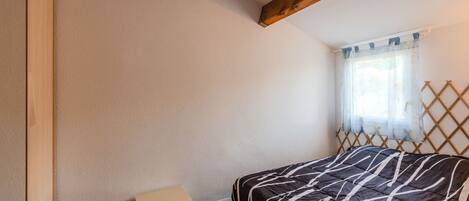 A bedroom with a bed, a small table, wooden beams on the ceiling, and a window with a curtain.