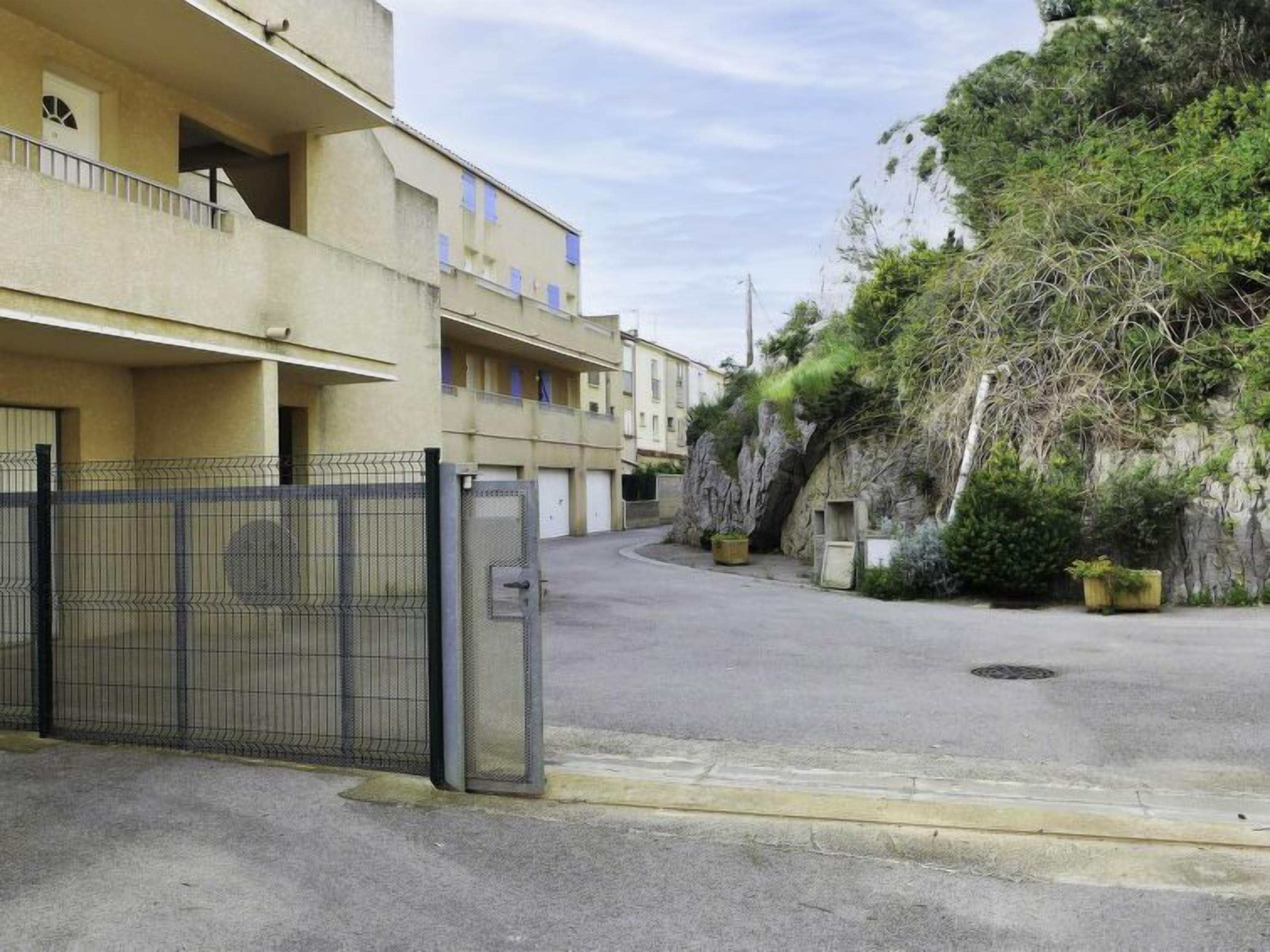 Ein durch einen Zaun gesicherter Parkbereich mit einem Gebäude, das Balkone besitzt, sowie eine felsige Böschung mit Vegetation.