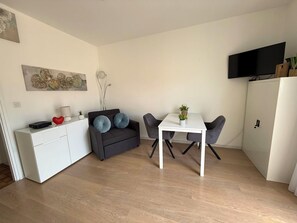A compact living space with a sofa, a dining table, a white cabinet, and a flat-screen TV mounted on the wall.
