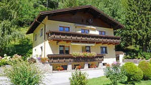 A two-story chalet with a balcony, surrounded by lush greenery and a mountainous backdrop.