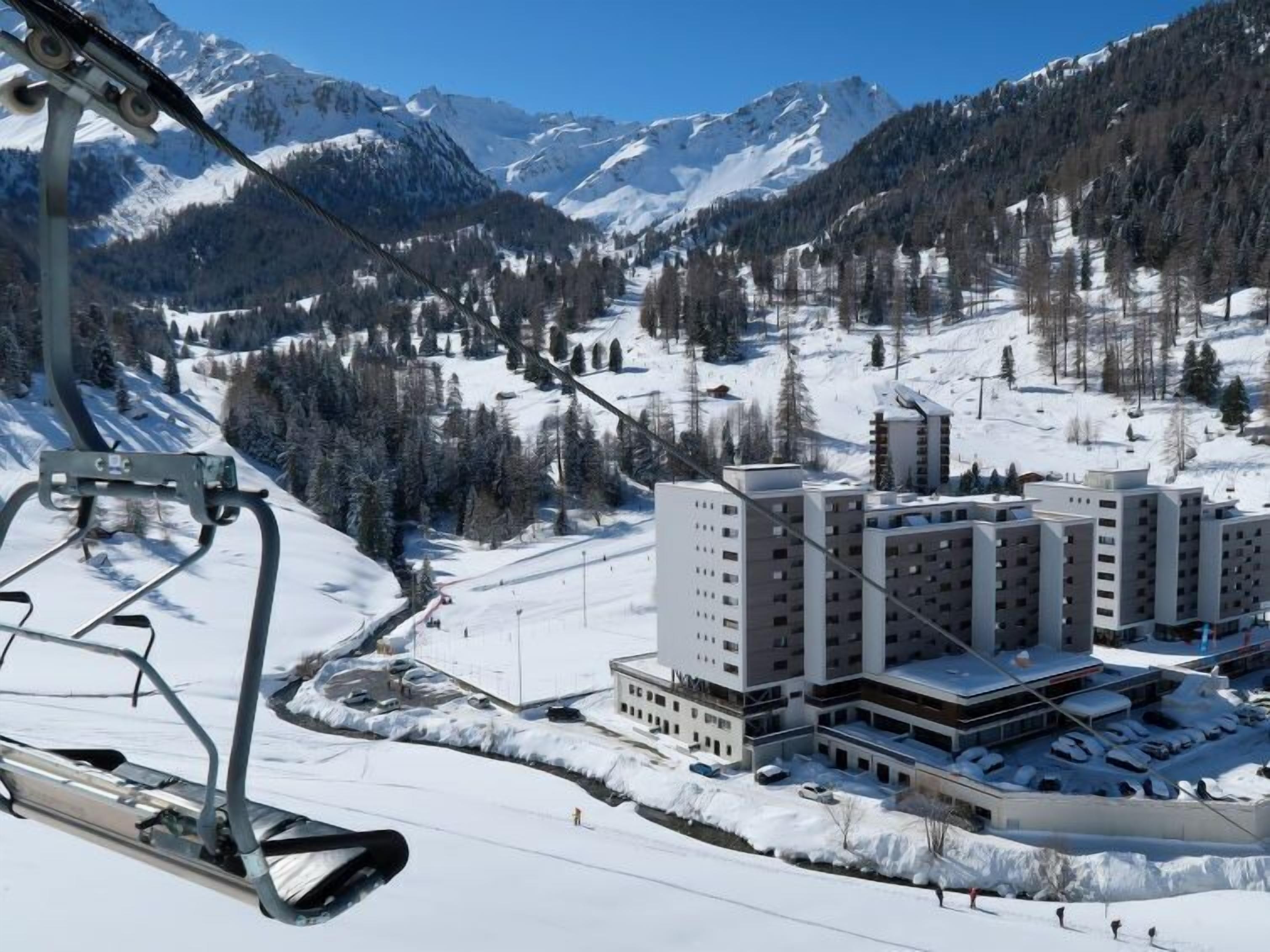 A ski lift with a view of a modern building complex surrounded by snow-covered mountains.