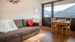 A cozy living room with a sofa, a red heart-shaped cushion, a dining table with chairs, and a large window offering a mountain view. - Pelvoux 102 by Interhome (Les Menuires)