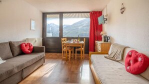 A cozy living room with a sofa, a dining table, and a view of mountains through a large window. - Pelvoux 102 by Interhome (Les Menuires)