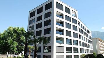 Un bâtiment moderne à plusieurs étages, doté d’une façade à motifs géométriques de fenêtres et de balcons, entouré de verdure et surplombant un ciel dégagé.