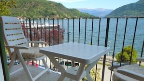 Un balcon avec une table et deux chaises, donnant sur un lac où l’on aperçoit des bateaux, avec des collines en arrière-plan.