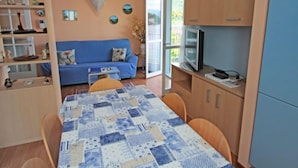 Un salon chaleureux avec un canapé bleu, une table basse recouverte d’une nappe à motifs et un téléviseur posé sur un meuble en bois.