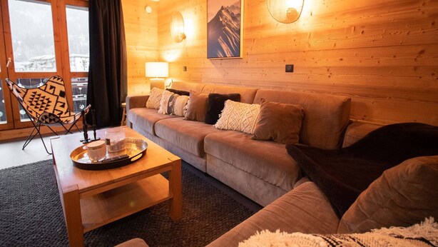 A cozy living room with a sofa, a coffee table, and a chair. There is a window with curtains, a wall-mounted lamp, and a framed picture. - 5-room duplex for 10 guests with balcony, Hameau de la Forêt (Valmorel)