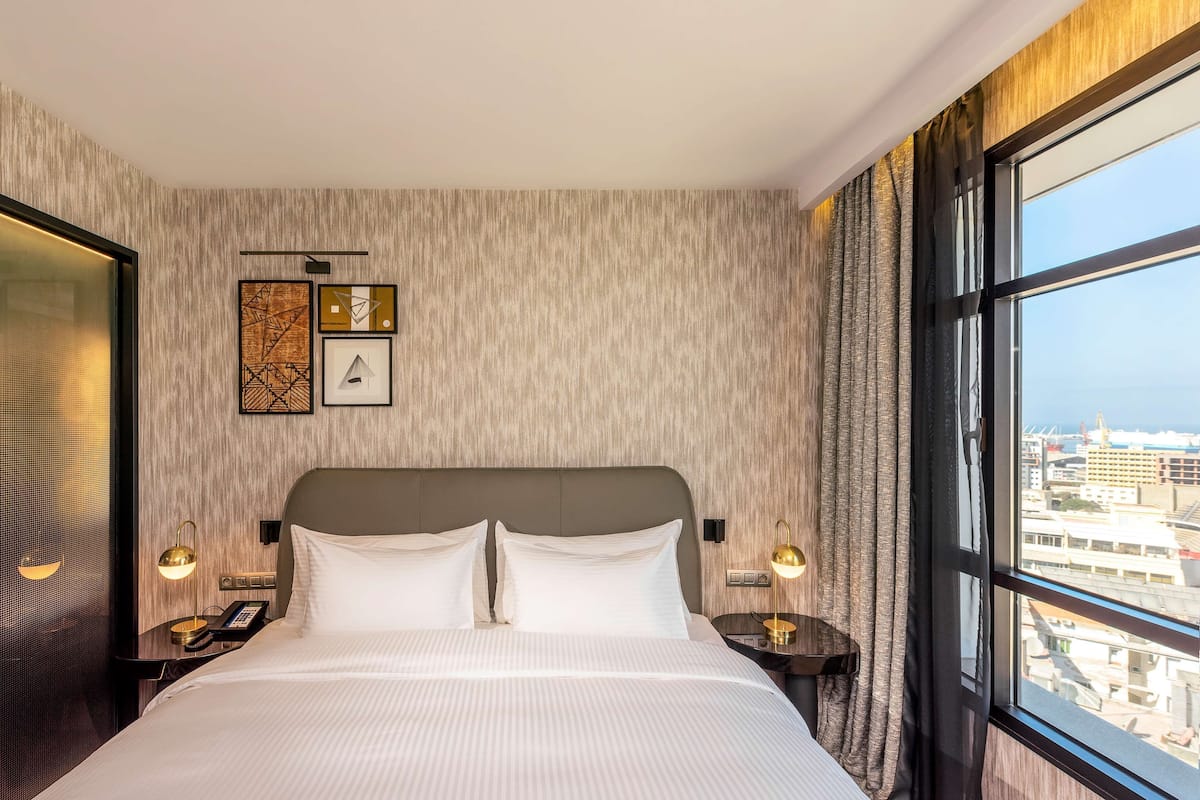A hotel room with a large bed, two bedside tables with lamps, a wall-mounted light fixture, and a view of the city through a large window.