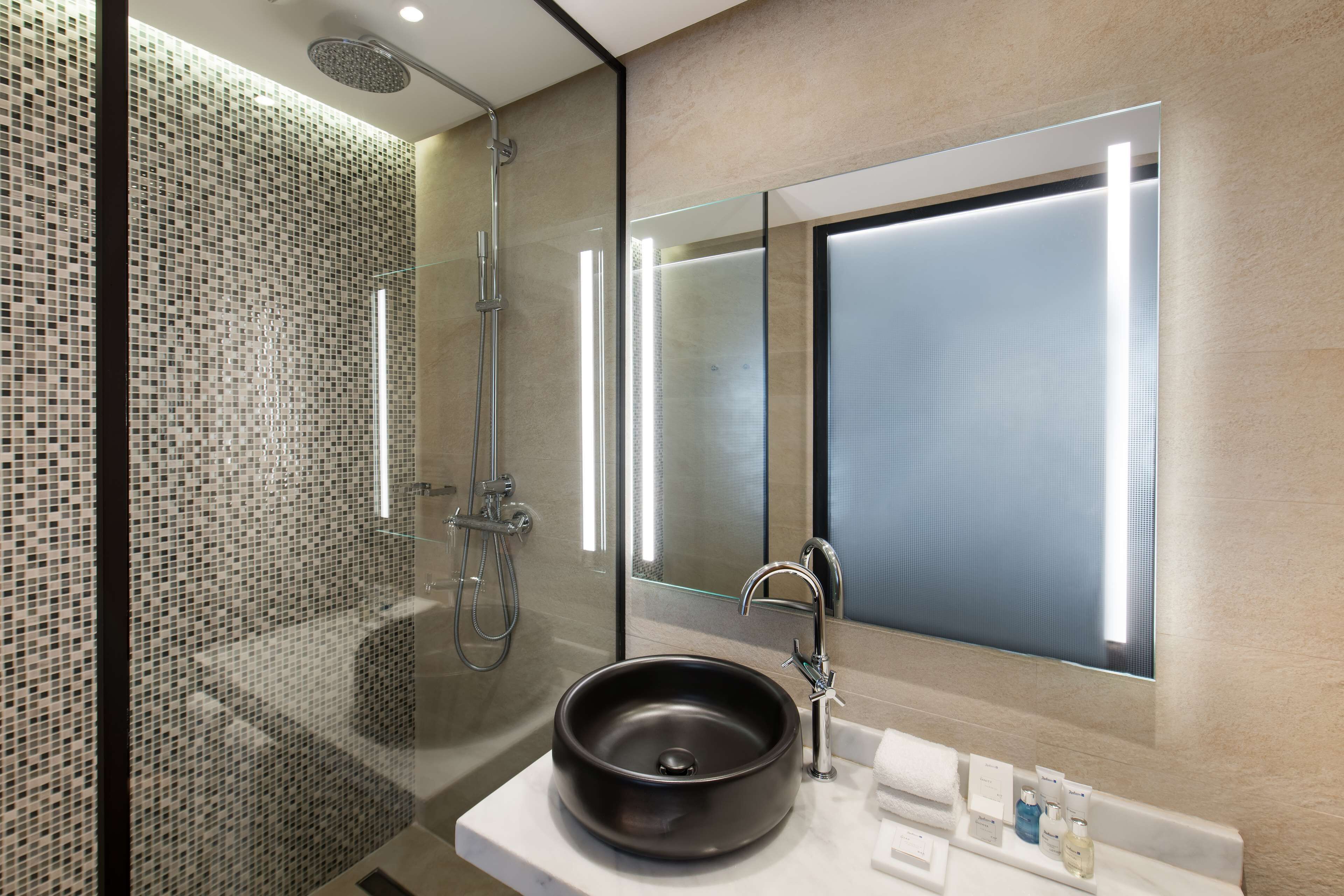 A modern bathroom with a glass-enclosed shower, a round black sink, and a large mirror with integrated lighting.