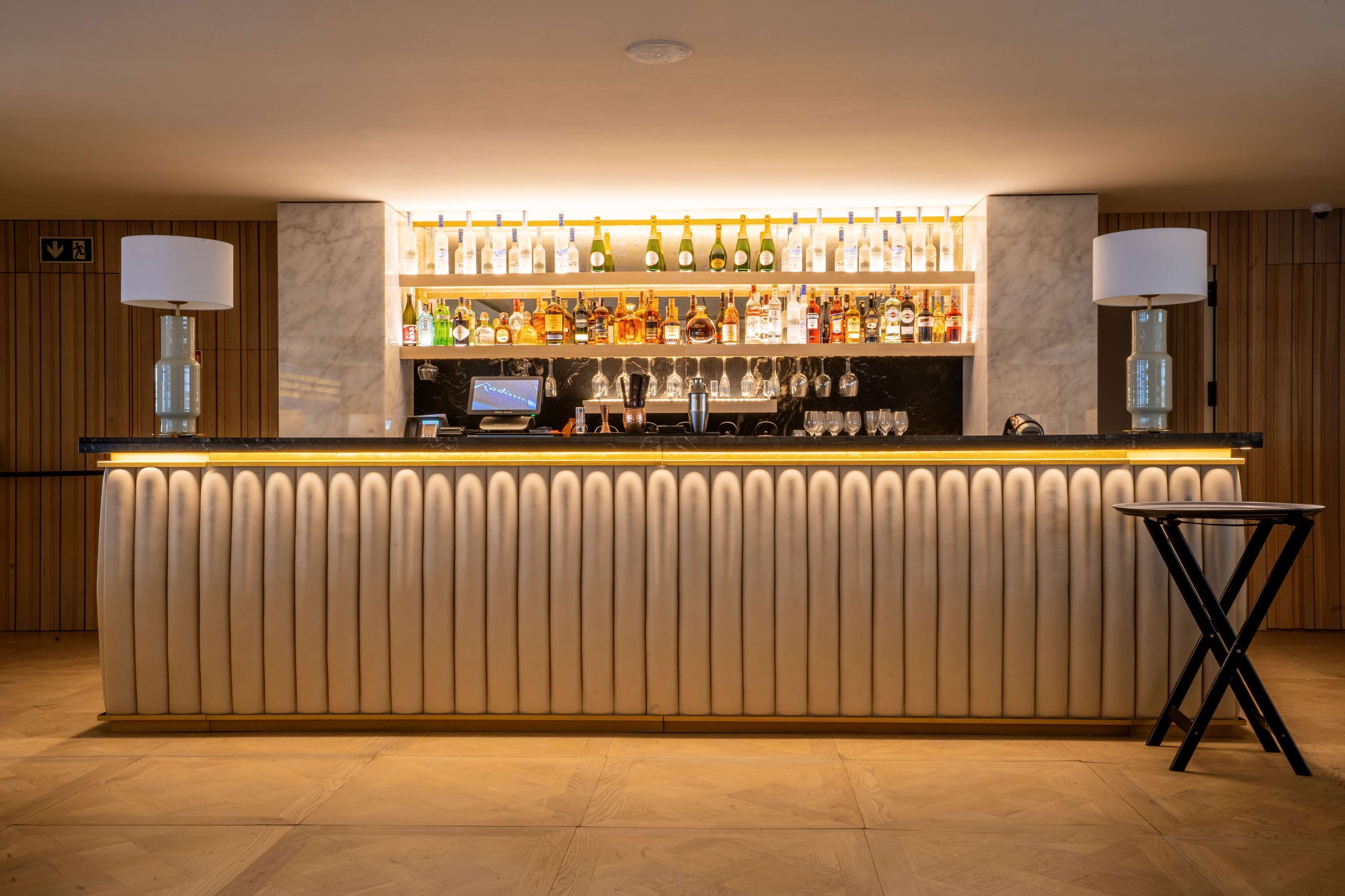Eine Bar mit einer Marmor-Theke, Regalen voller Spirituosenflaschen und zwei Lampen.