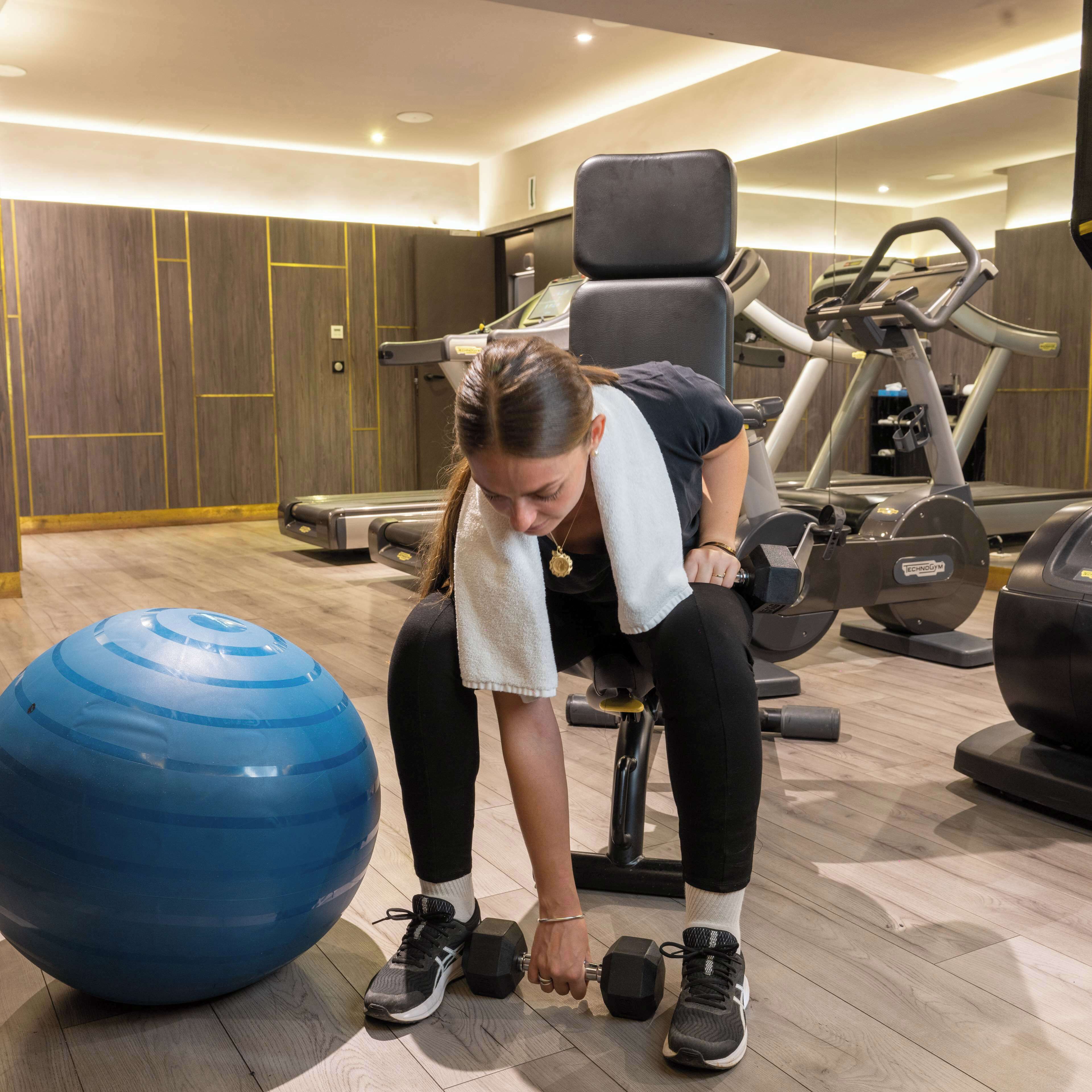 A gym with exercise equipment, a blue exercise ball, and a person lifting weights.
