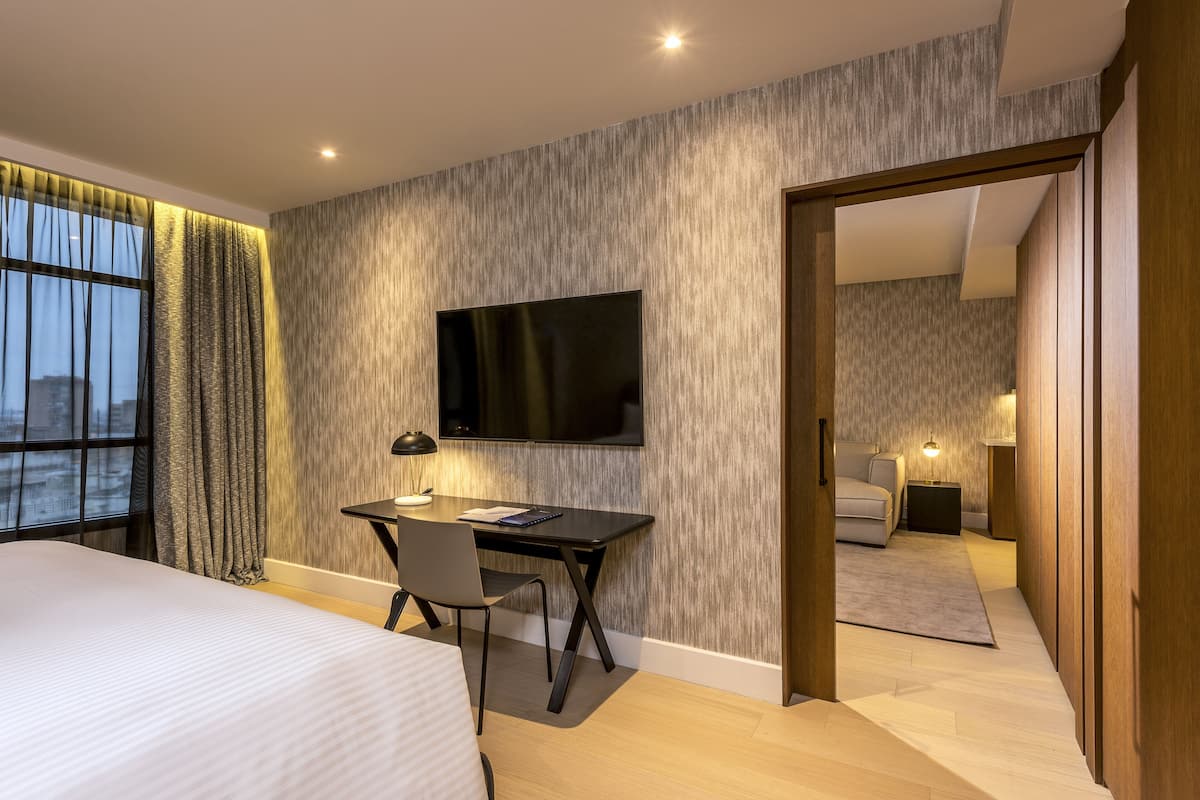 A modern hotel room with a large bed, a desk with a chair, a flat-screen TV mounted on the wall, and a view of the city through the window.
