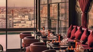 Un restaurante con vista a la ciudad, mesas con cojines rojos y un ventanal grande.