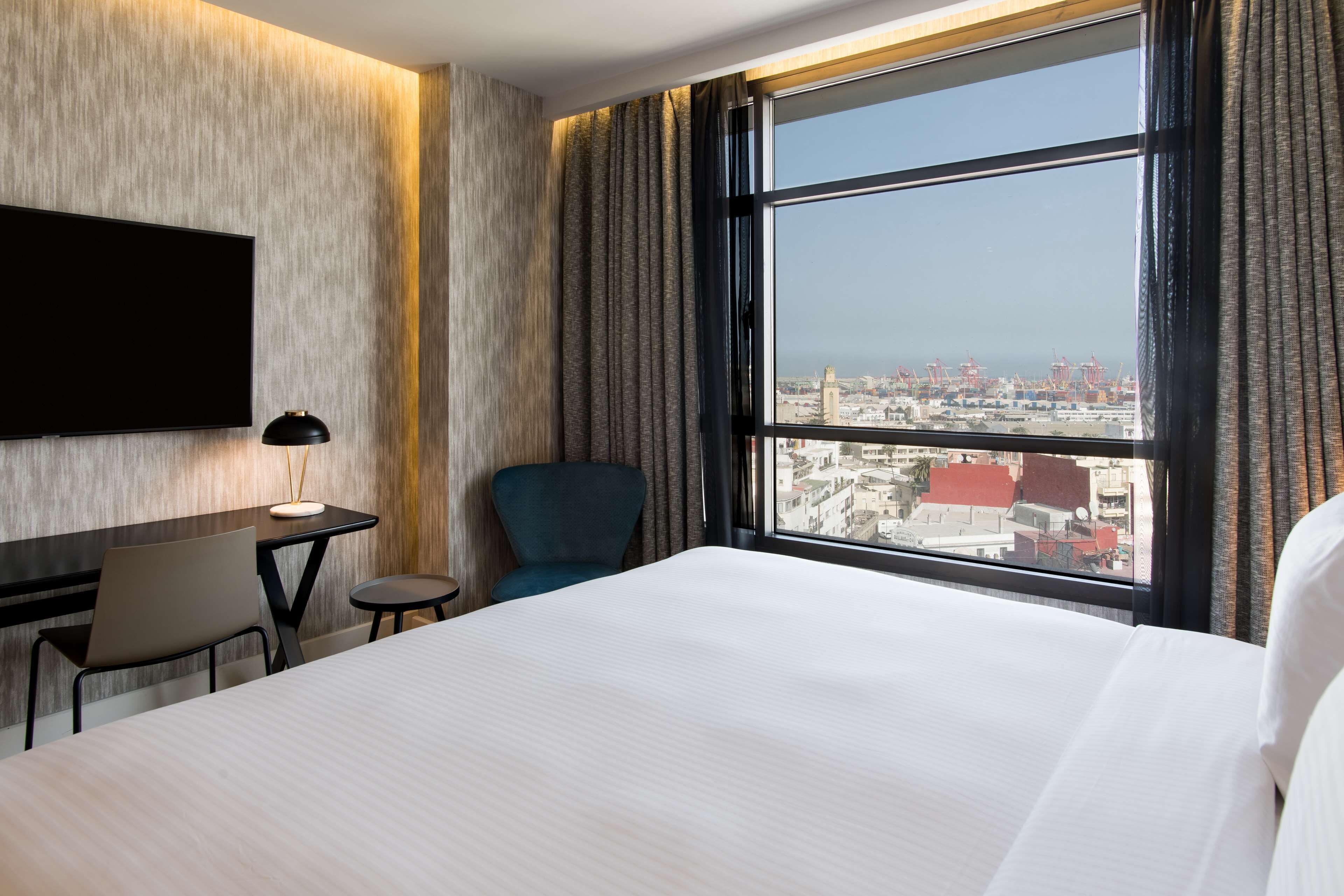 Camera d'albergo con un letto grande, una scrivania con sedia, una TV e vista sulla città da una finestra ampia.