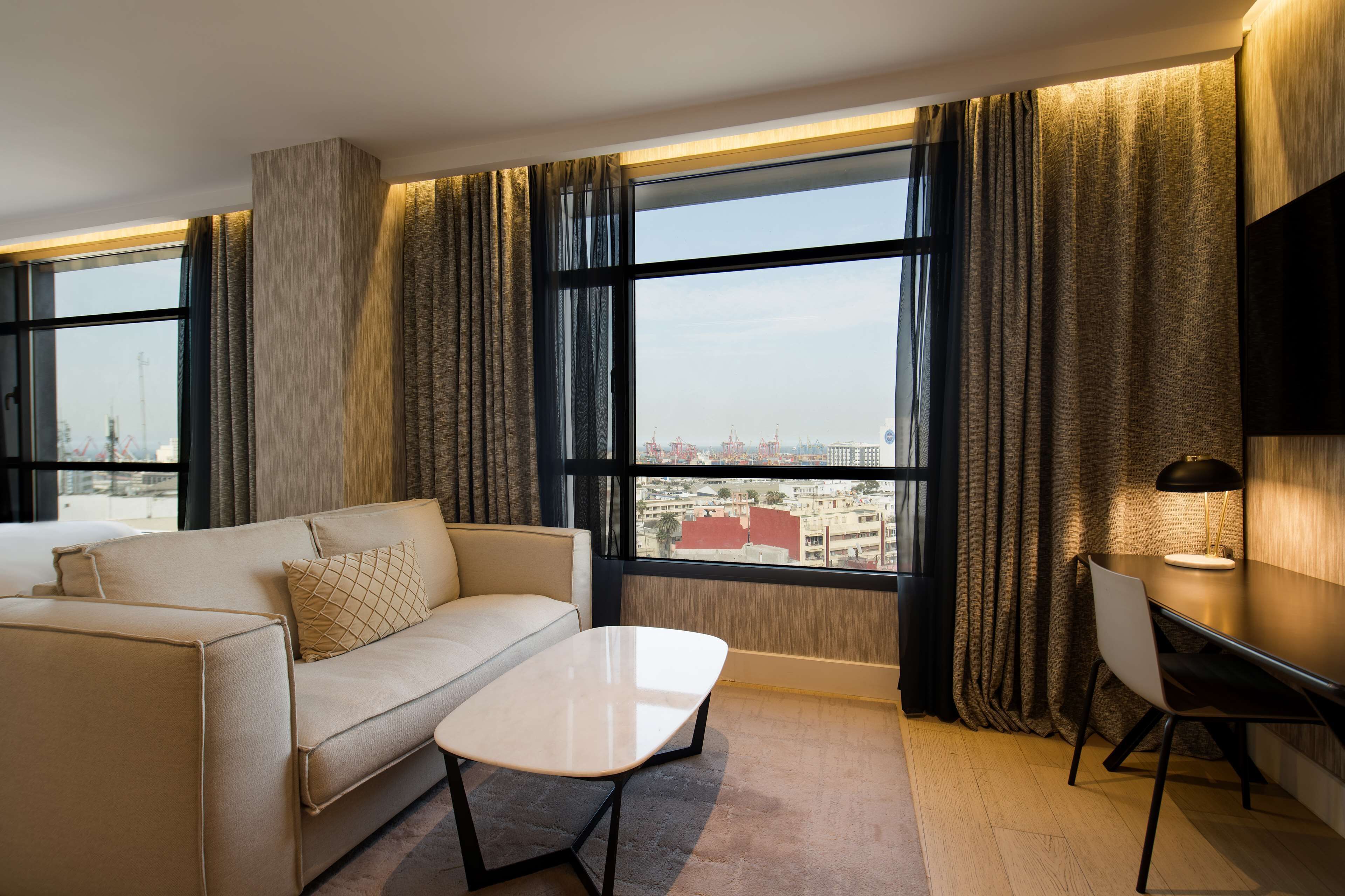 A modern hotel room with a sofa, a small table, a desk with a lamp, and large windows offering a city view.