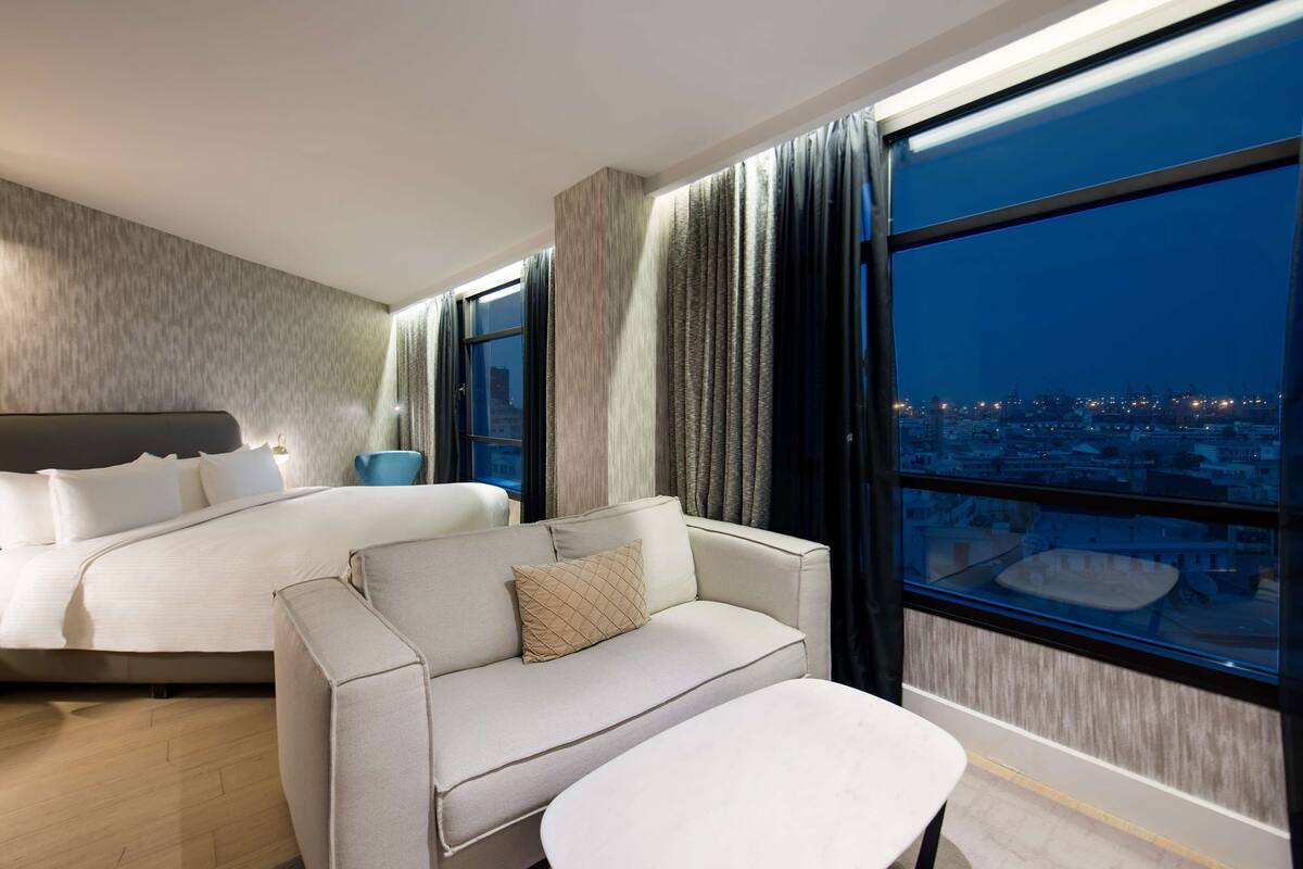 A modern hotel room with a large bed, a sofa, a small table, and a city view through large windows.