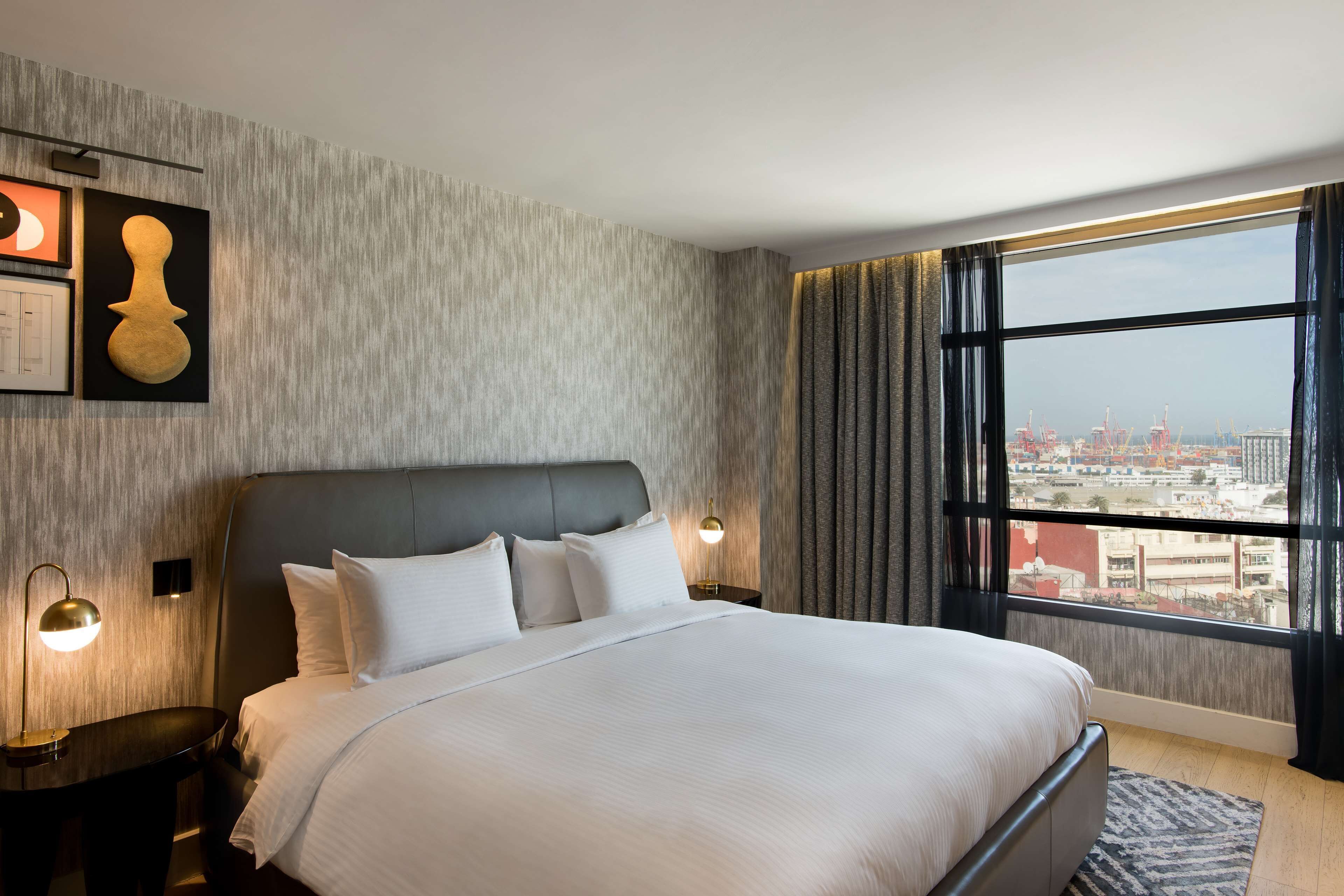 A modern hotel room with a large bed, a city view, and a textured wall.