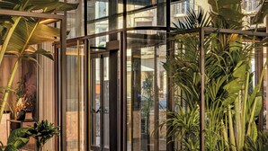 Un vestíbulo moderno con un ascensor de vidrio, plantas en macetas grandes y una zona de estar con sofá y reposapiés.