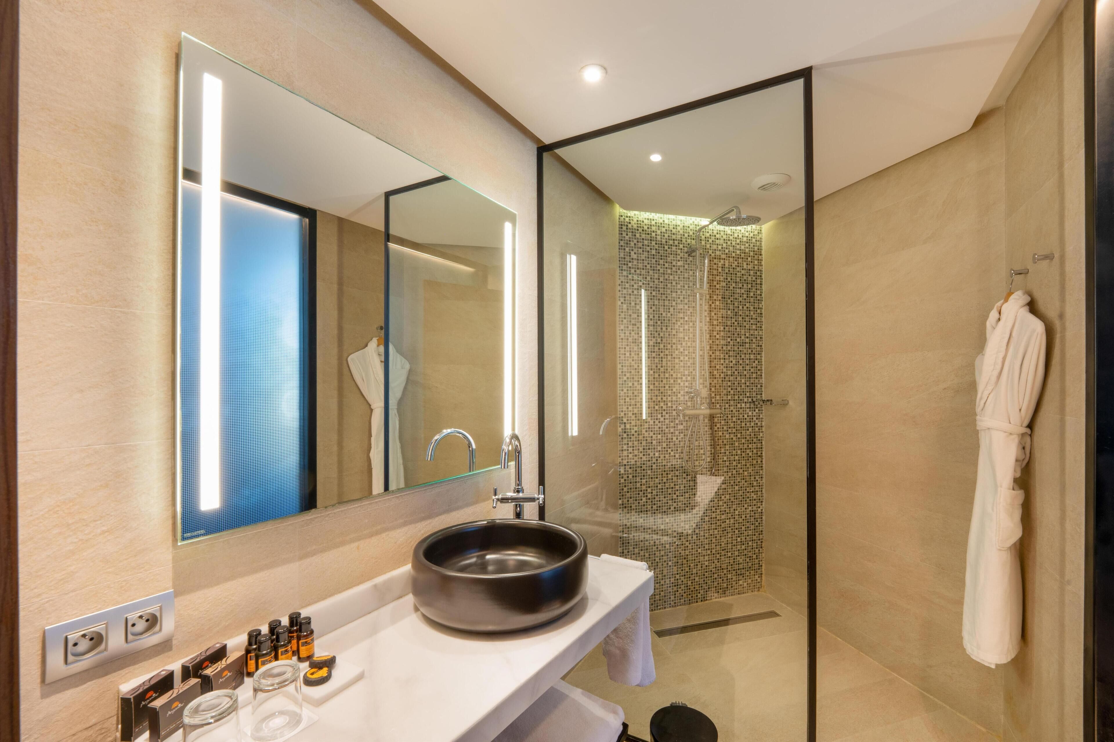 A modern bathroom with a large mirror, a round sink, a glass shower enclosure, and a white robe hanging on the wall.