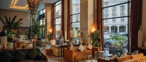 A spacious lobby with a green sofa, brown armchairs, and a marble coffee table. Large windows offer views of the city.