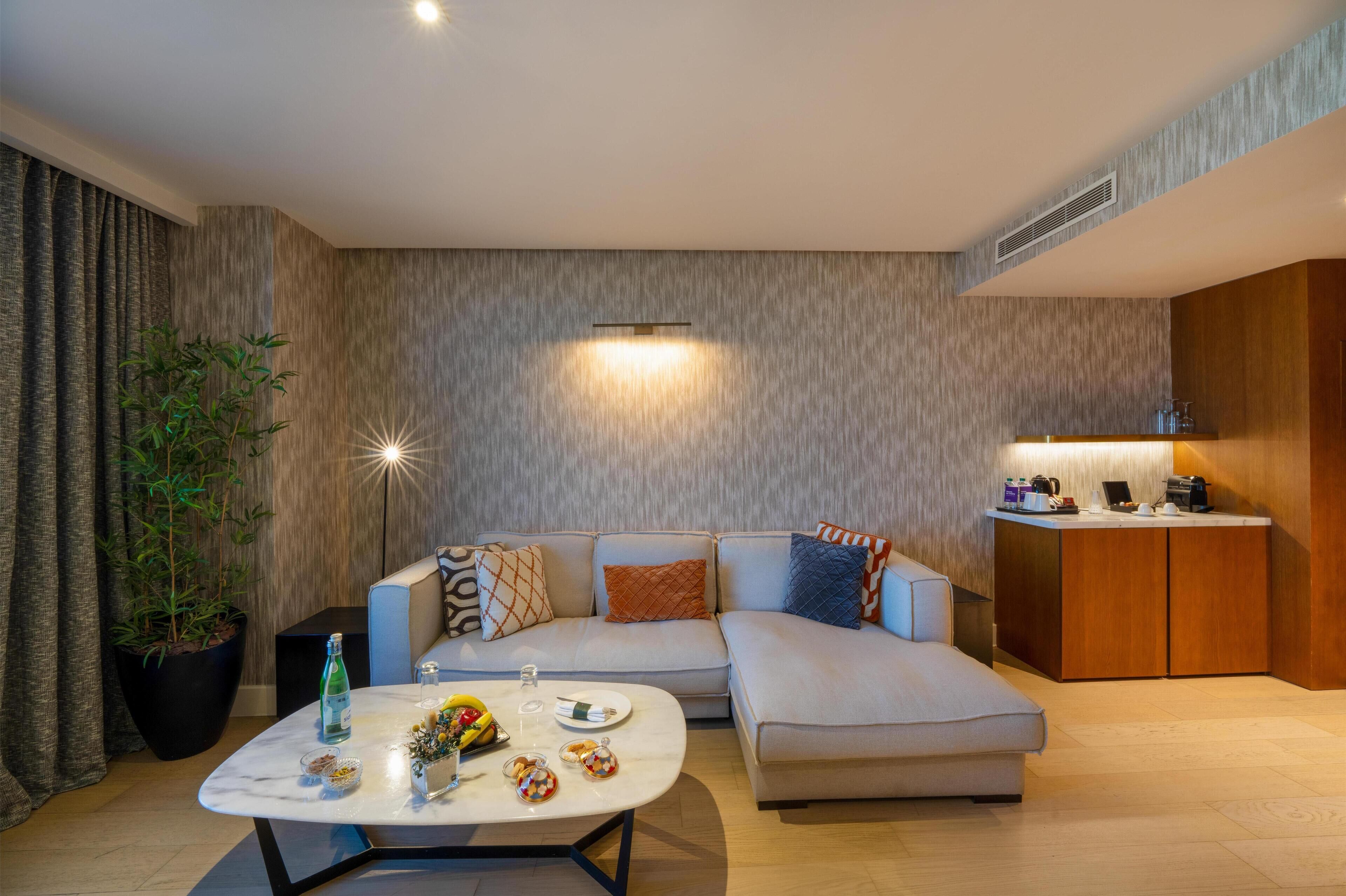 Ein modernes Wohnzimmer mit Ecksofa, Couchtisch mit Dekoration, Zimmerpflanze und integrierter Küchenzeile mit Arbeitsplatte und Schränken.