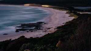 Un paysage côtier avec une plage, une côte rocheuse et des collines à l’horizon.