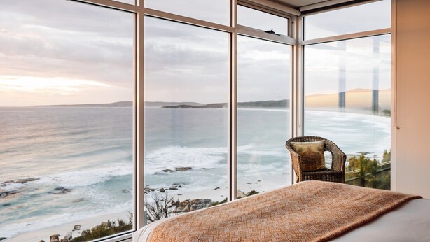 Une chambre avec une grande fenêtre donnant sur la côte, une chaise en osier et un plaid à motifs.