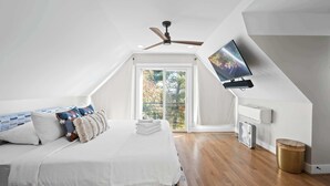 Un dormitorio moderno con techo inclinado, cama con ropa blanca, ventilador de techo, televisor de pantalla plana montado en la pared y ventana con vista a árboles.