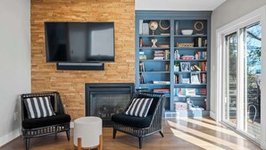 Una sala de estar con un televisor montado en una pared de ladrillo, dos sillones de mimbre negro con cojines a rayas, un reposapiés blanco y una estantería llena de libros y objetos decorativos.