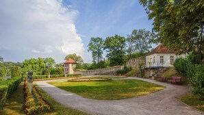 Um jardim com paisagismo, uma alameda pavimentada, uma torre de pedra e um edifício com telhado de telhas vermelhas.