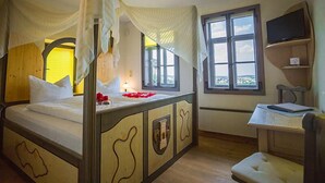 Quarto com cama de dossel, escrivaninha com cadeira e televisão na parede.