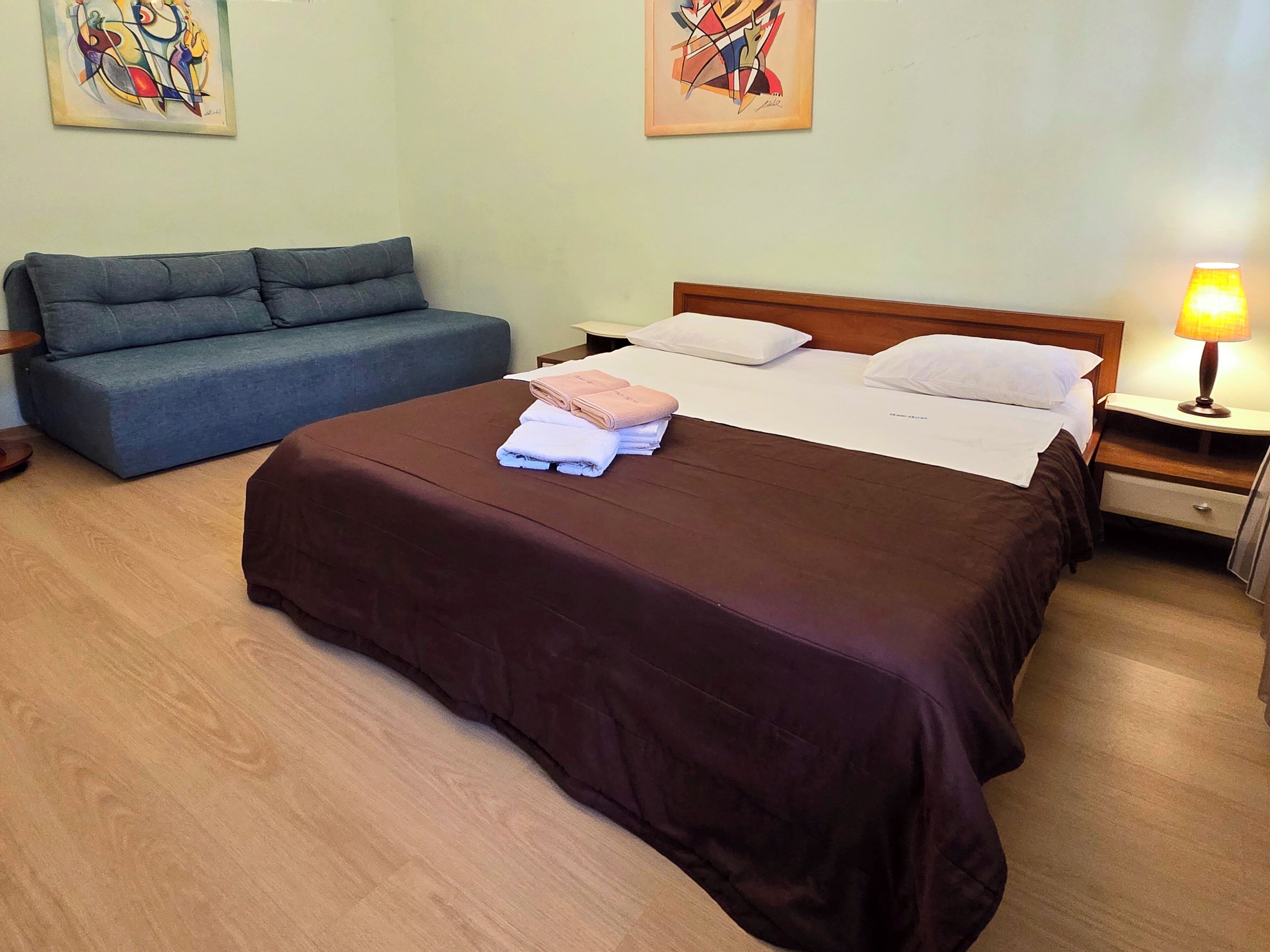 A hotel room with a bed, a sofa, a nightstand with a lamp, and artwork on the walls.