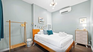 Quarto moderno com cama de madeira, roupa de cama branca, mesa de cabeceira com abajur e ar condicionado fixado na parede.