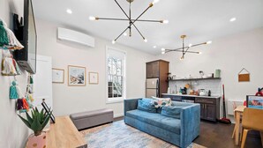 Uma sala moderna com um sofá azul, uma mesa de centro de madeira e um tapete estampado. Há uma área de cozinha com armários e uma geladeira ao fundo.