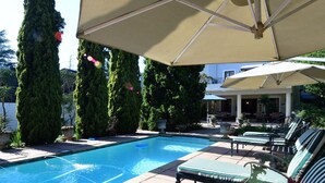 A swimming pool with lounge chairs and umbrellas, surrounded by tall trees and a building in the background. - Highgrove Guesthouse (Sandton)