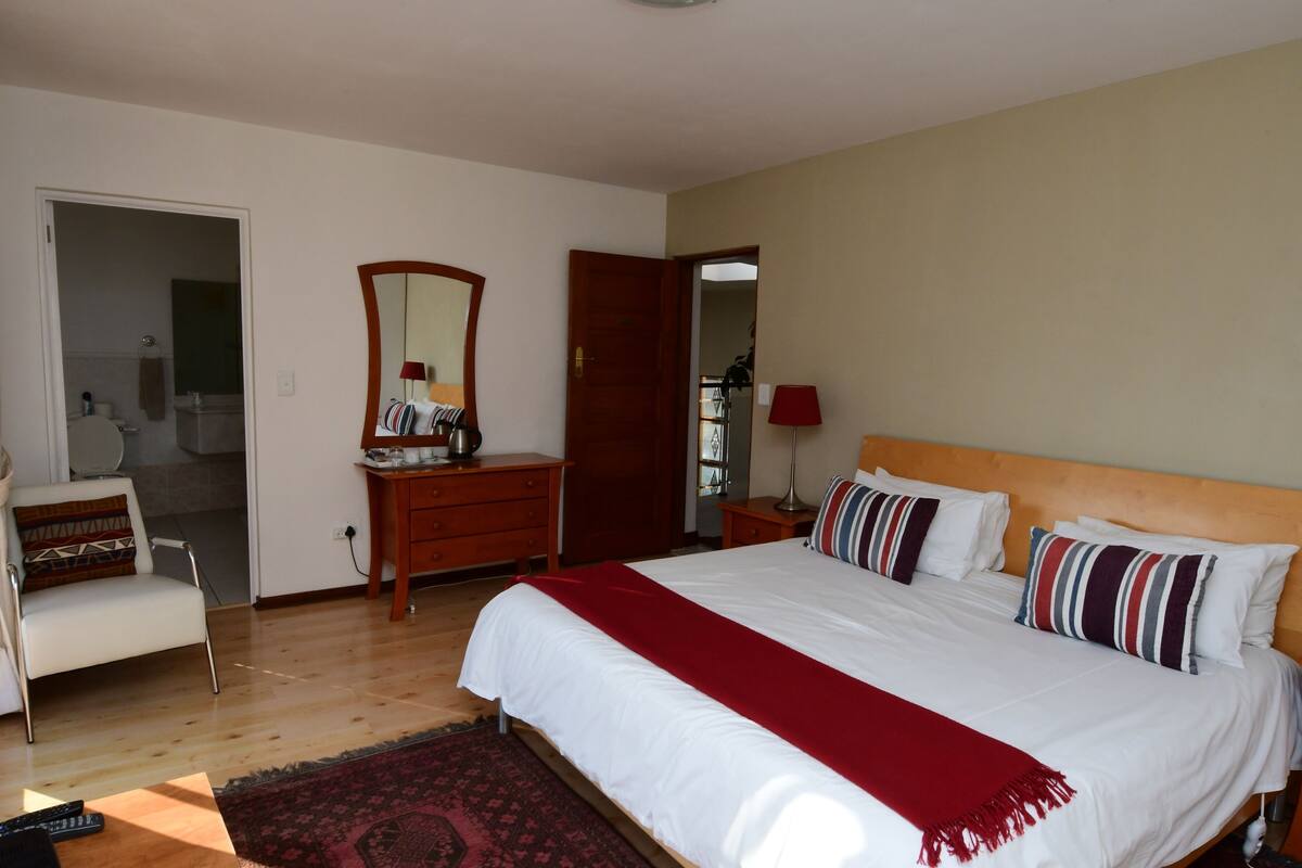 Een slaapkamer met een groot bed, een houten dressoir, een stoel, een spiegel en een deur naar een badkamer.