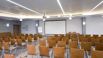 Un salón de conferencias con filas de sillas de madera, una pantalla blanca grande y un proyector.