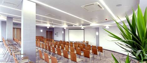Un salón de conferencias con filas de sillas de madera y una pantalla grande al frente.