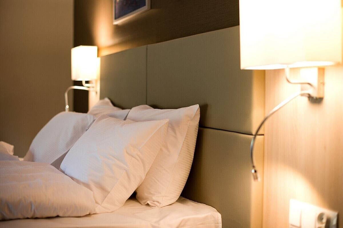 Ein ordentlich gemachtes Bett mit weißen Leinen und zwei an der Wand befestigten Lampen, die Licht spenden.