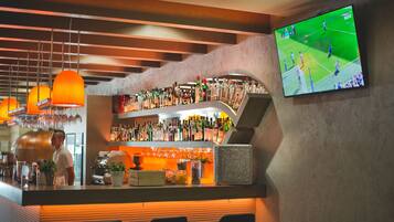 Een bar met flessen drank achter de bar, een televisie waarop een sportwedstrijd te zien is, en een barman achter de toog.