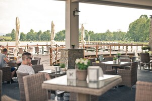 Restaurante ao ar livre com mobiliário de vime, mesas e guarda-sóis, com vista para um corpo d'água.