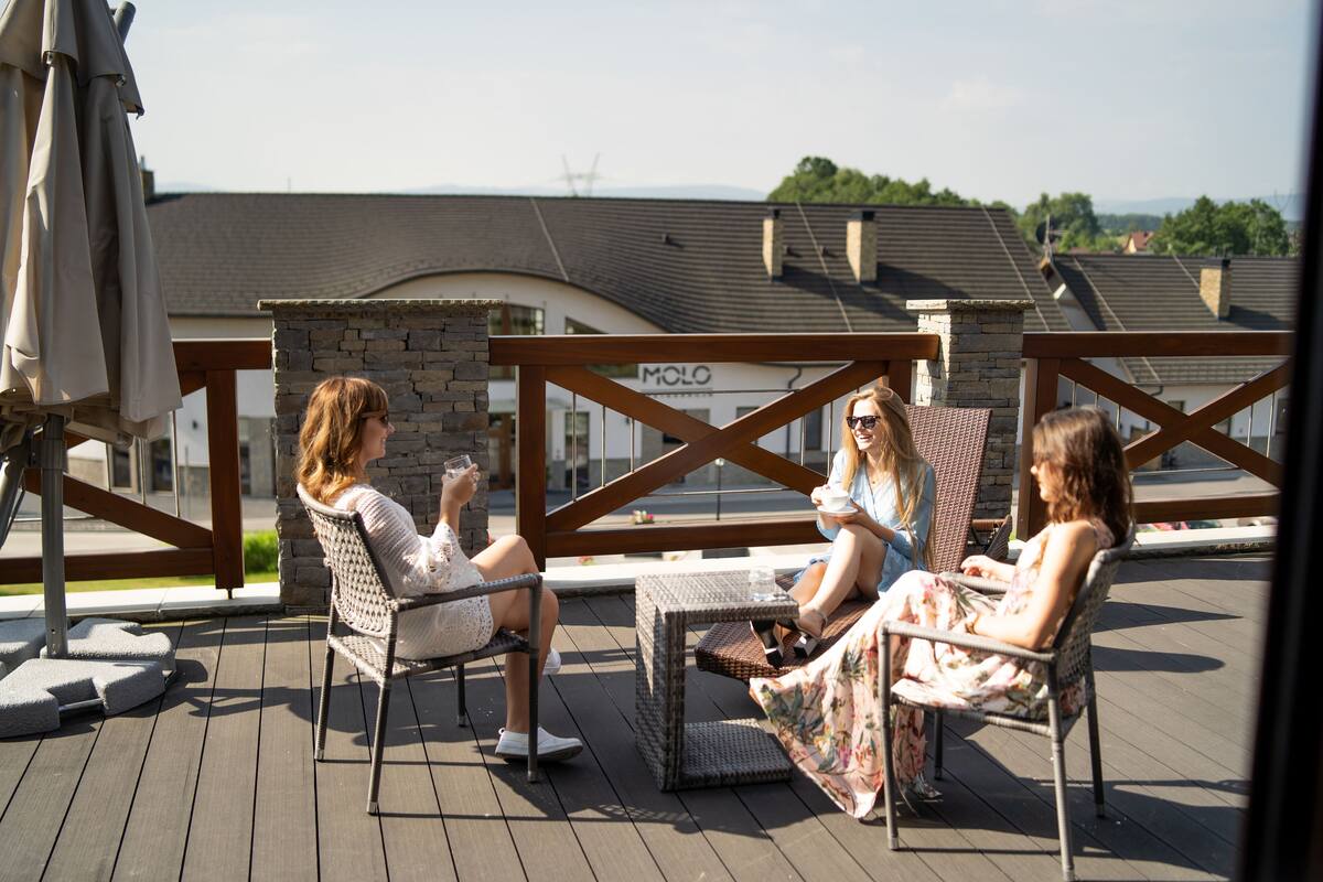 Drei Personen sitzen auf Korbstühlen auf einer Holzterrasse mit Steinmauern und einem Metallgeländer.