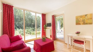 A room with a red sofa, a white table with a red tablecloth, and a white chair. There is a large window with red curtains, a radiator, and a TV on a stand. - Agriturismo Rossolampone (San Bernardino Verbano)